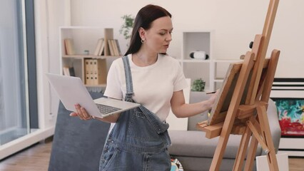 Wall Mural - Female professional painter having video conference using portable computer in comfy flat with amazing panoramic view. Attractive female in casual outfit having online art class with beginners.