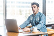 © khosrork - Portrait of amazed astonished surprised man freelancer in blue jeans shirt working on laptop, having video conference, looking at display with big eyes. Indoor shot near big window, cafe background.
