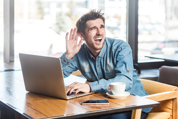 Portrait of bearded man freelancer in blue jeans shirt working on laptop, having problems with microphone speaker, keeps hand near ear, trying to hear. Indoor shot near big window, cafe background.