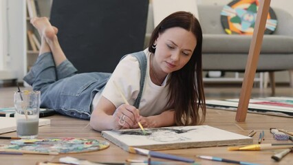 Wall Mural - Young attractive woman dressed in white t-shirt and denim jumpsuit lying on floor of cozy apartment and posing on camera with paintbrush. Talented artist drawing picture while staying at home.