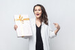 Portrait of surprised astonished brunette woman holding pointing gift box, looking at camera with big eyes and open mouth, wearing striped jacket. Indoor studio shot isolated on gray background.