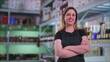 © Marco - Smiling Female Supermarket Employee with Arms Crossed Standing in Aisle, Joyful Expression Next to Product Inventory