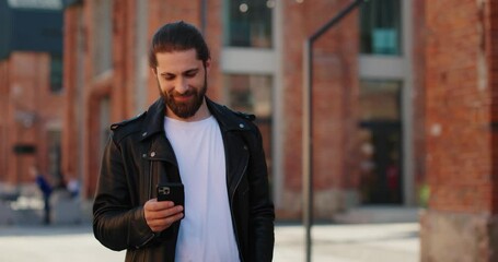 Stylish man in leather jacket walking down the street while using smartphone. 
