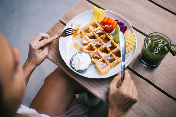 Cropped female customer visiting local cafeteria for eating crispy fresh baked wafer using cutlery at wooden table with match cocktail, unrecognizable woman with knife and fork eating Belgian waffle