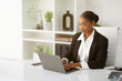 Beautiful black businesswoman in formal wear working with laptop computer at desk in office interior, copy space
