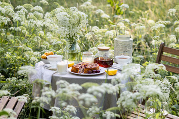 Tea time with sweet homemade cherry cake and herbal beverage in summer garden outdoors. Table covered with linen tablecloth, white porcelain cups. English style