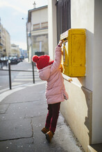Adorable Preschooler Girl Putting Letter In Yellow Post Box On A Street Of Paris