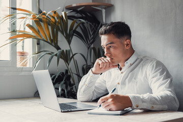 Wall Mural - Thoughtful businessman touching chin, pondering ideas or strategy, sitting at wooden work desk with laptop, freelancer working on online project, student preparing for exam at home.