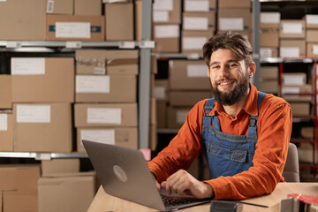 Wall Mural - Warehouse worker with laptop entering inventory data to database, smiling and looking at camera