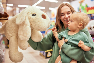 Happy young mother with her cute daughter walking around toy store in shopping mall. Mom and little blonde baby girl looking at a big soft toy and having fun. Funny weekend, happy childhood.