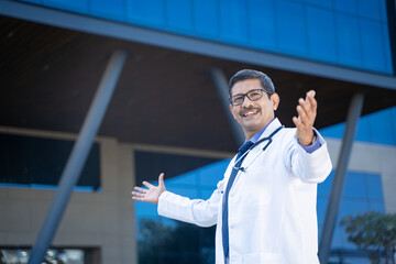 Wall Mural - Confident Indian doctor spreading hand and giving happy expression out of hospital.