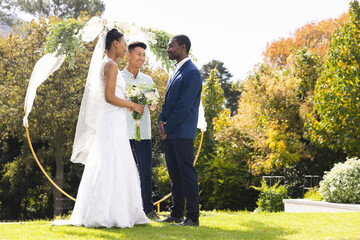 Happy diverse male officiant, bride and groom at outdoor wedding in sunny garden, copy space