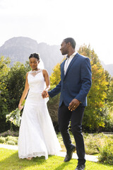 Wall Mural - Happy african american bride and groom holding hands, walking in sunny garden, copy space