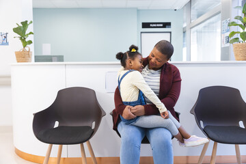Happy african american mother and daughter sitting in waiting room at hospital, copy space