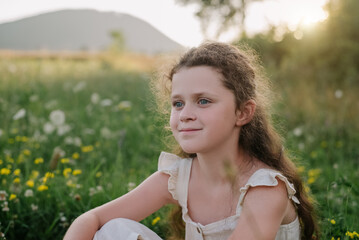 Portrait of happy preteen girl sitting on green grass outdoor, enjoying beautiful summer nature during warm sunset or sunrise. People emotions, travel, adventure, childhood and wanderlust concept