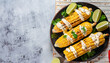 Elotes, Grilled Mexican Street Corn, charred cobs are covered in creamy mayonnaise, seasoned with chili powder and sprinkled with cheese, cilantro, and a spritz of lime juice, flat lay, free space