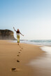 Barefoot white woman running in the beach on a clear and sunny day. Girl on focus on first view and cliff on the background. She wears green pants and white shirt and it takes place in a sandy beach