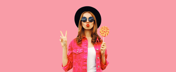 Wall Mural - Portrait of happy young woman holding lollipop and blowing her lips sends air kiss wearing pink jacket, black round hat on pink background