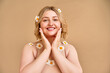 © Velista production - Portrait of a beautiful young woman with a round face and curvaceous forms touching her cheeks and smiling. Daisies in the hair, on the shoulders and on the chest. Body positive. Skin care.