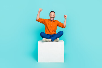 Photo of ecstatic man with white gray beard wear stylish shirt sit on cube raising fists up win bet isolated on teal color background