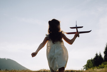 Silhouette of little girl kid run with airplane on background amazing mountains during warm summer sunset. Dream freedom concept. Playful child runs on green field holding in hands toy aircraft