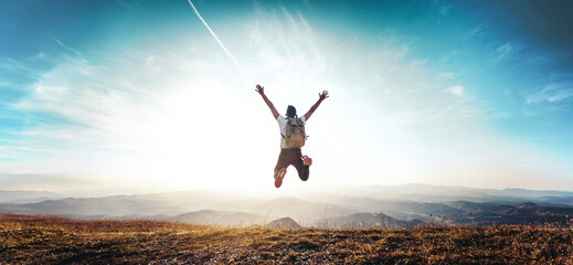 Wall Mural - Happy man with arms up jumping on the top of the mountain - Successful hiker celebrating success on the cliff - Life style concept with young male climbing in the forest pathway