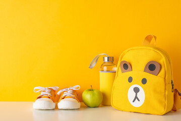Cheerful school time for small kids concept. Side-view photo of orange backpack and sneakers, water bottle and apple for snack on yellow isolated backdrop with copy space for text placement or advert
