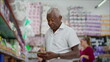 © Marco - One senior black man shopping for products standing at grocery store aisle browsing items while holding and looking at phone