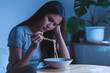 © KMPZZZ - End of month. Alone depressed asian young student woman, girl using chopsticks eating instant ramen, noodles, cheap food and no money for dinner in bedroom in overtime night, late time at home.