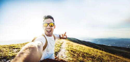 Wall Mural - Happy man with backpack and sunglasses taking selfie picture on top of the mountain - Cheerful hiker climbing the cliff outdoors - Travel blogger looking at camera - Pov view