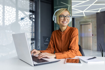 Young beautiful woman working on laptop inside office at workplace, smiling businesswoman listening to online podcast, audio book and music, female worker studying listening to training course.