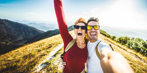 Wall Mural - Cheerful couple of hikers taking selfie on top of the mountain - Millennial guy and girl enjoying summertime day out laughing at camera together - Millenial travelers standing on nature background