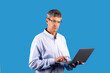 Mature businessman holding laptop frowning typing on blue studio background