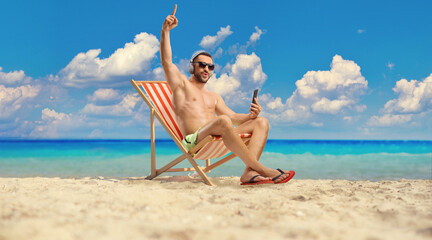 Wall Mural - Young man in swimwear sitting on a beach chair and enjoying with headphones