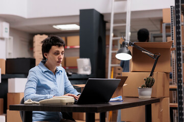 Wall Mural - Storehouse employee looking at cargo stock on laptop computer while checking shipping detalies before start preparing customers packages. Worker working at warehouse delivery department