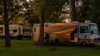 Rv and trailers parked at campsites amongst tall trees in early morning with grass and sunrise light shadows