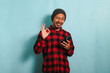 Satisfied Young Asian man with a beanie hat and a red plaid flannel shirt is winking his eyes while holding a mobile phone and showing an OK gesture, isolated on a blue background