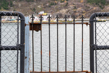 A Wire Railing Fence And Wrought Iron Gate Overlook The Water. The Fencing Is Used To Protect People From Falling Into The Ocean. There's A Pier In The Distance With Small Fishing Boats Moored. 