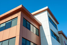 The Roofs Of A Modern Commercial Building Under Blue Sky And White Clouds. The Exterior Of The New Building Is Rusty, Orange, And Cream In Color Metal Composite Panels With Black Glass Windows. 