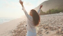 Young Positive Indian Woman Stands With Back To Camera And Raises Hands Up Enjoying Warm Evening Sun And Not Wanting To Leave Beach Dressed In White Clothes Posing On Seashore. Travel Lifestyle