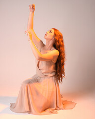 Poster - Full length portrait of beautiful brunette  female model wearing dress with golden back light lighting. Kneeling pose with gestural arm pose.  isolated on studio background.