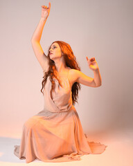 Poster - Full length portrait of beautiful brunette  female model wearing dress with golden back light lighting. Kneeling pose with gestural arm pose.  isolated on studio background.