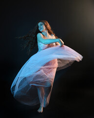 Wall Mural - Full length  portrait of beautiful brunette woman dancer, wearing ethereal gown, dancing with flowing fabric.  isolated on dark studio background with cinematic colourful lighting.