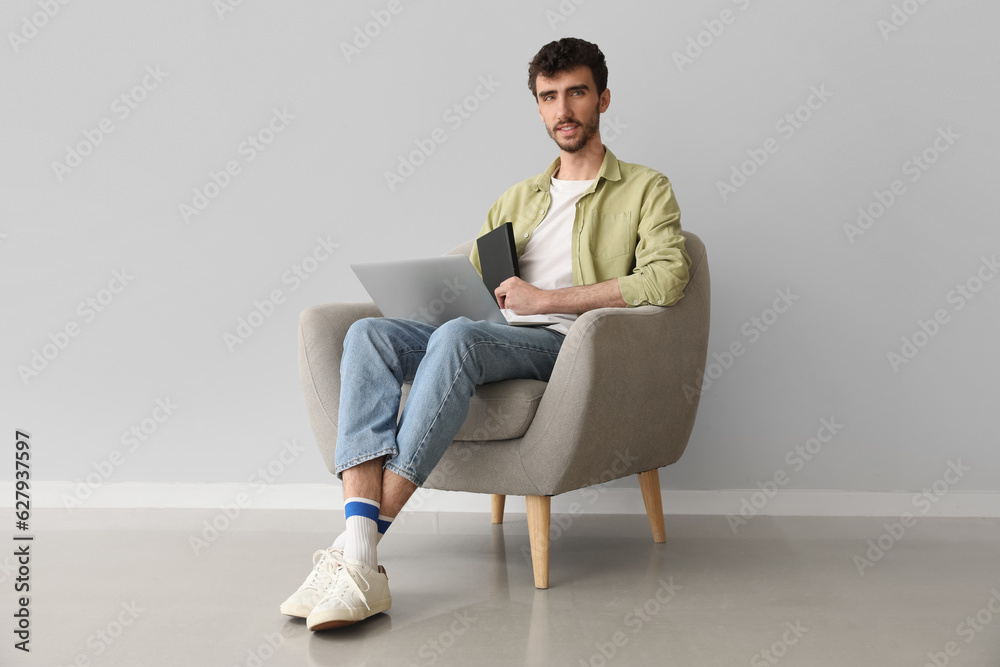 Young man working with laptop and notebook in armchair near light wall