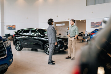 Wall Mural - Remote view of doubtful male customer buying car in showroom talking to sales agent in business suit looking at auto indoors in spacious dealership. Concept of choosing and buying auto at showroom.