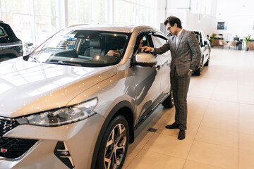 Wall Mural - Wide shot of successful customer male talking to sales representative in motor showroom sitting inside brand new car holding steering wheel. Concept of buying new auto in dealership office.