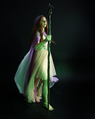 Wall Mural - portrait of beautiful brunette woman wearing  a gown with purple fantasy cloak holding a wooden wizard staff, isolated on dark studio background with cinematic colourful lighting.
