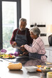 Happy senior biracial couple using tablet preparing vegetables in kitchen at home, copy space