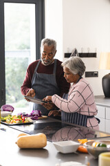 Happy senior biracial couple using tablet preparing vegetables in kitchen at home, copy space