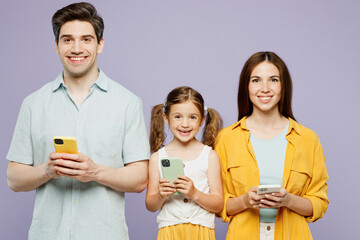 Wall Mural - Young happy parents mom dad with child kid daughter girl 6 years old wear blue yellow casual clothes hold in hand use mobile cell phone chatting isolated on plain purple background Family day concept